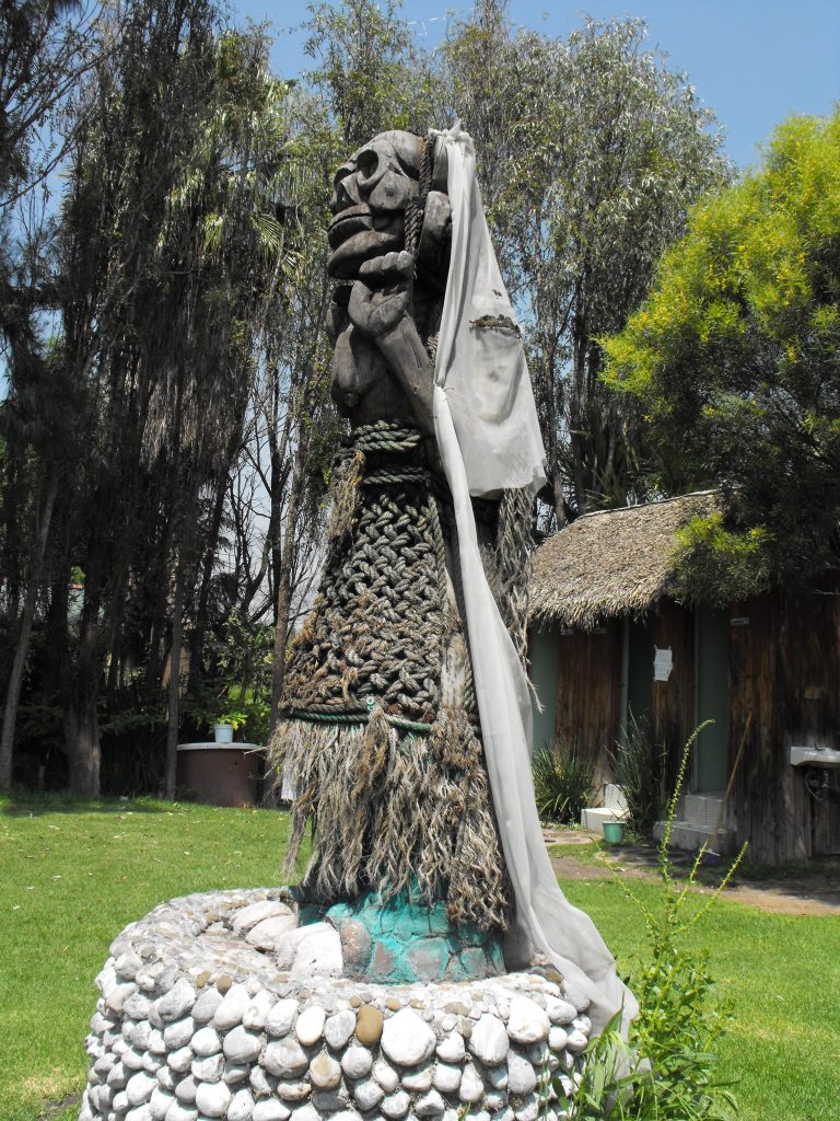 Picture in color on a sunny day of a statue of La Llorona in Xochimilco, Mexico. There is a green grass lawn and a stone pedestal holds the statue of La Llorona. La Llorona is an Indigenous weeping woman figure. She wears a veil and a long dark gown. "Wood carved in the shape of a woman called "La llorona", typical of Mexican culture, with a white veil on a stone base, located on the island of "la llorona" in the canals of Xochimilco, Mexico."