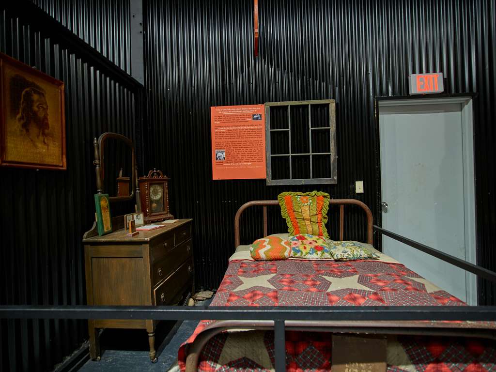 photo of a museum display with a made bed and furnitures in the foreground and a false window and exit door in the background.
