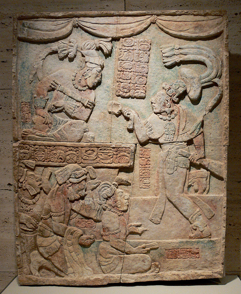 Limestone carving of a maya king sitting on top facing a standing subject with three kneeling figures at the bottom.