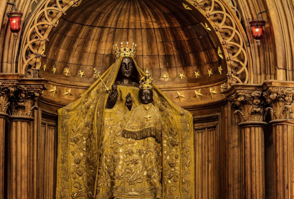 Photo of a statue in an apse (arched alcove) with columns of a woman with black skin holding a child with black skin, both wearing ornate gold robes and bejeweled crowns. The woman is holding a sceptre in her right hand and what appears to be an apple.