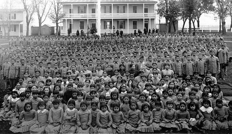 Black and white photo of rows of about 300 Native American children all in similar dress, the girls sitting in front and four times as many boys standing behind them, all in front of a white, two-story house