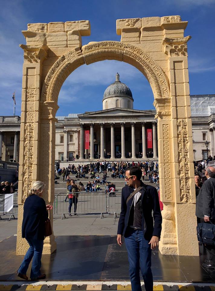 Photo of a sand-colored arch framing a building in the background with two people standing near it Sand-colored arc about 15 feet tall framing the collonaded National Gallery building in London with large crowd on its steps.