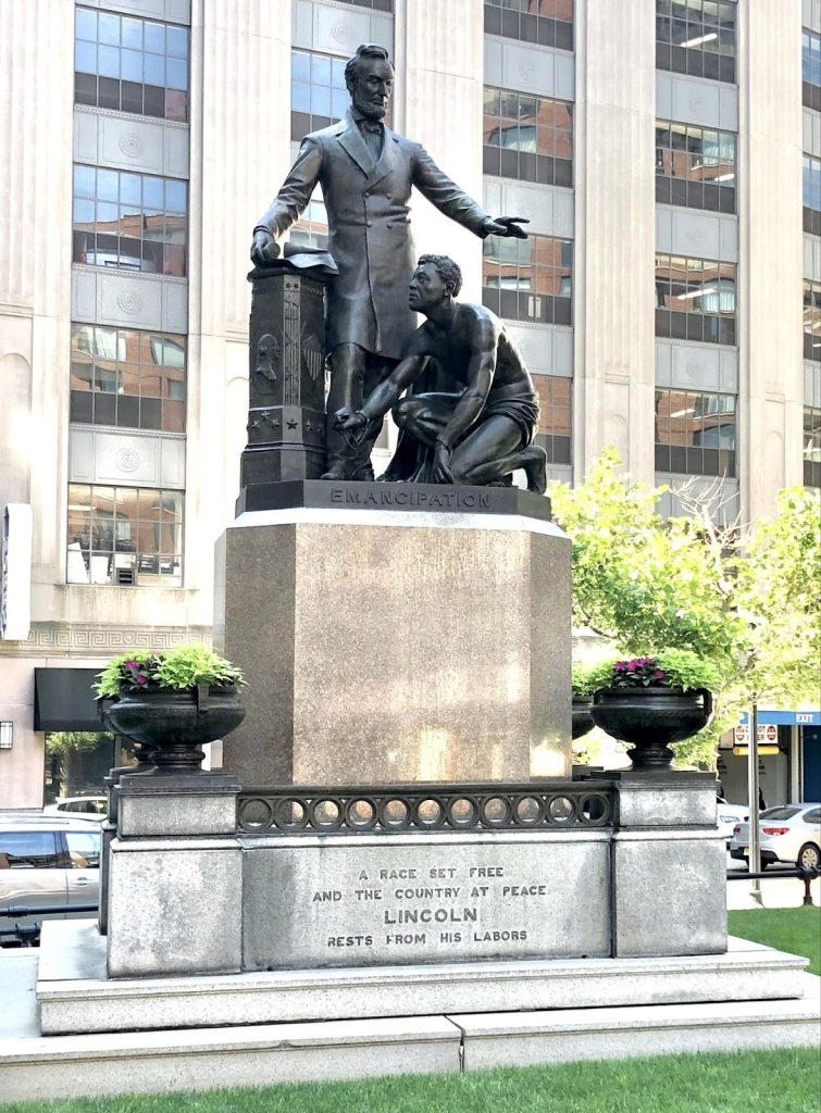 Large bronze statue on pedastal labeled Emanc.ipation, of a shirtless freed slave kneeling before President Lincoln, with his left arm outstretched and his right hand resting on the Emancipation Proclamation.