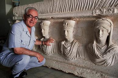 70-year-old man with glasses kneels down next to a sarcophagus from Palmyra dating back to the first century, with the busts of three people on the side shown.