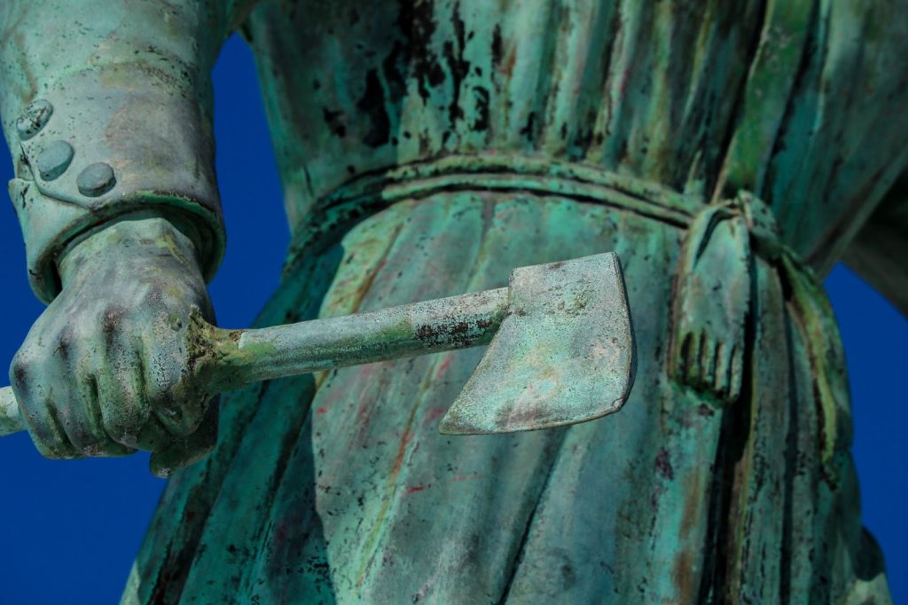 Close-up photo of a green-tinted copper statue of a woman in a robe holding a hatchet in her right hand.