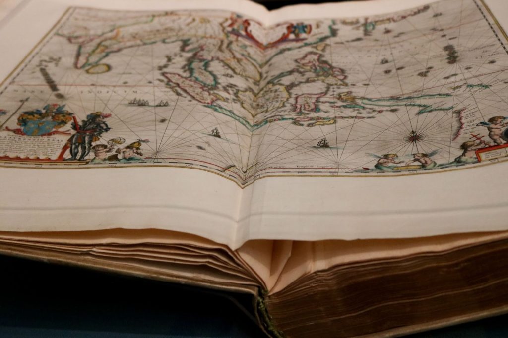 Photo of a book opened to a full page map with a landmass in the center and an illustration of an European explorer in the lower left corner and decorative cherubs along the lower border.