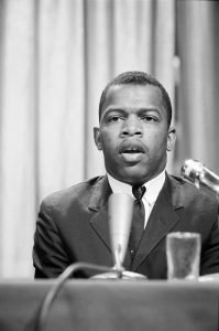 Black and white photo of a Black man in a suit and tie sitting in front of two microphones