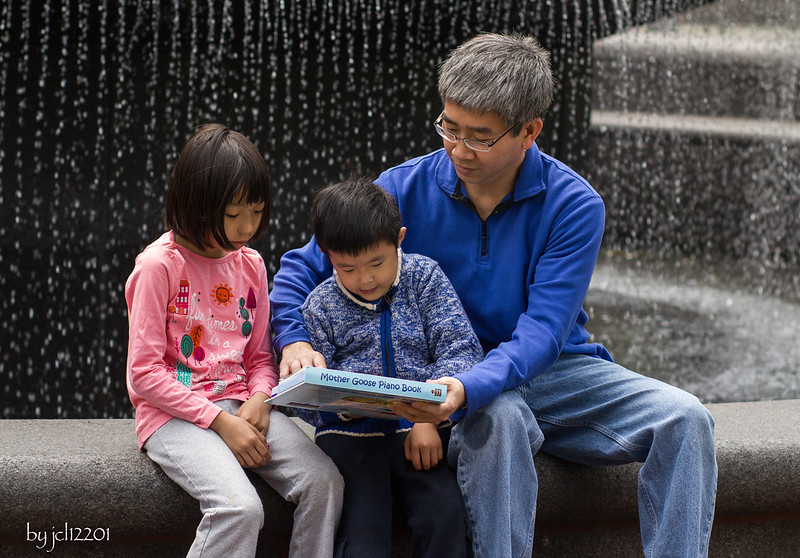 A parent and two children are sitting at a fountain. The parent holds the book in front of one of the children while the other looks on.