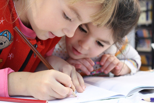 Photo of two young girls focus on writing with a pencil on a notebook.