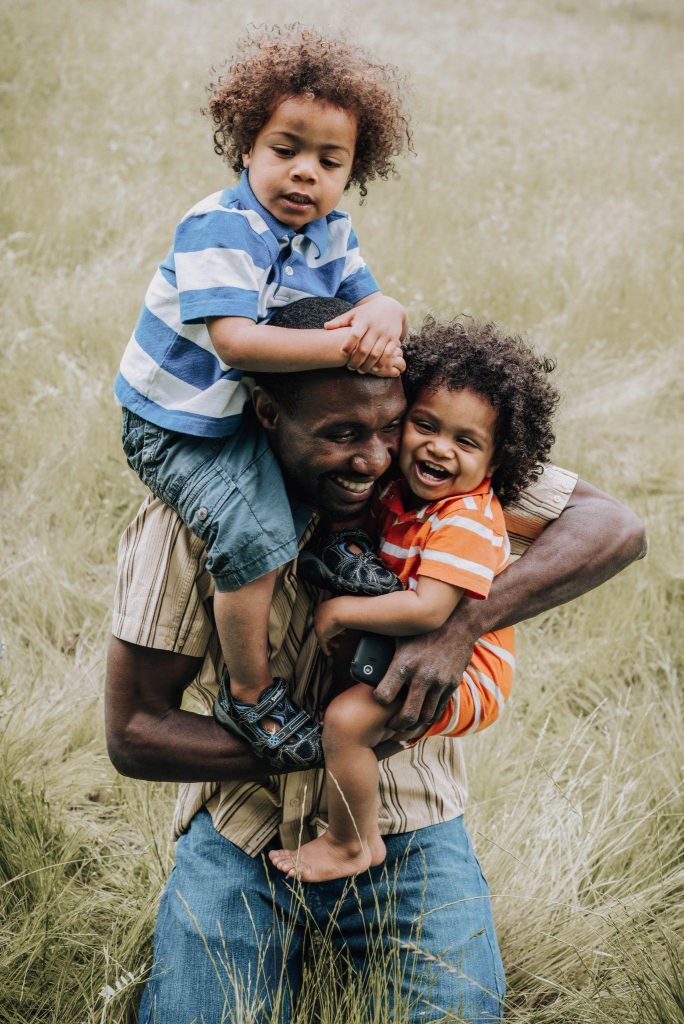 image of a father is laughing and smiling. He is holding one of his children on his right shoulder and the other in his arm. Both children are also laughing and smiling.
