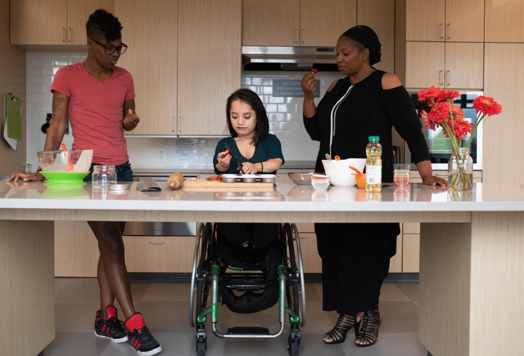 Image of three disabled people of color (a Black non-binary person on the left, a South Asian person with a wheelchair in the middle, and a Black woman on the right) at a kitchen counter with open space underneath. The person in the middle chops strawberries while the other two each sample a strawberry.