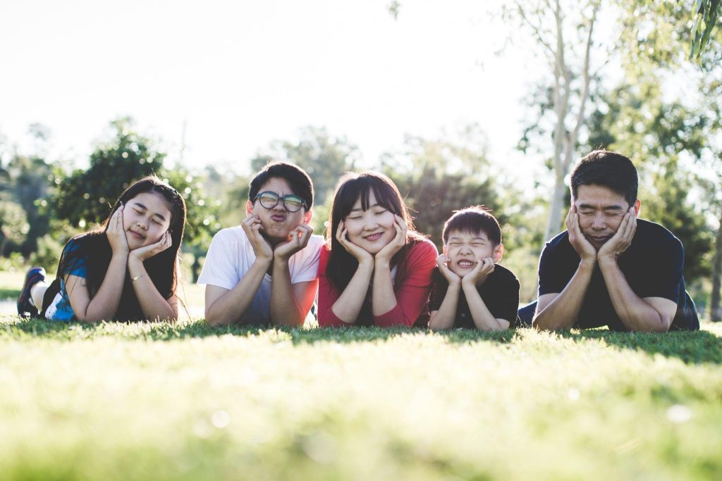 Image of a family of 5 lies on the grass with their hands holding their faces. They are making funny faces and smiling.