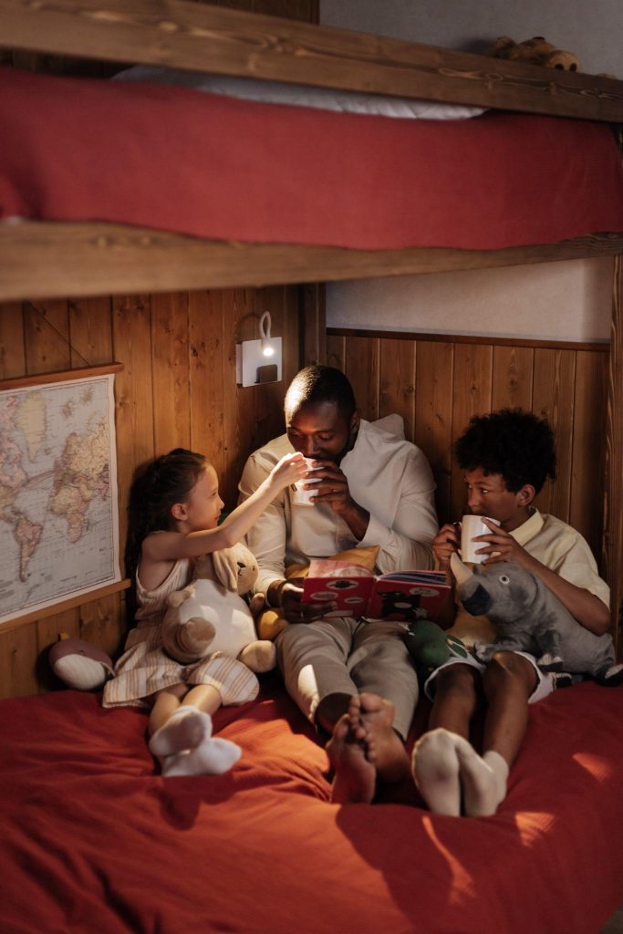 Image of a father sits in bed with his two children. They are drinking tea and reading a bed time story.