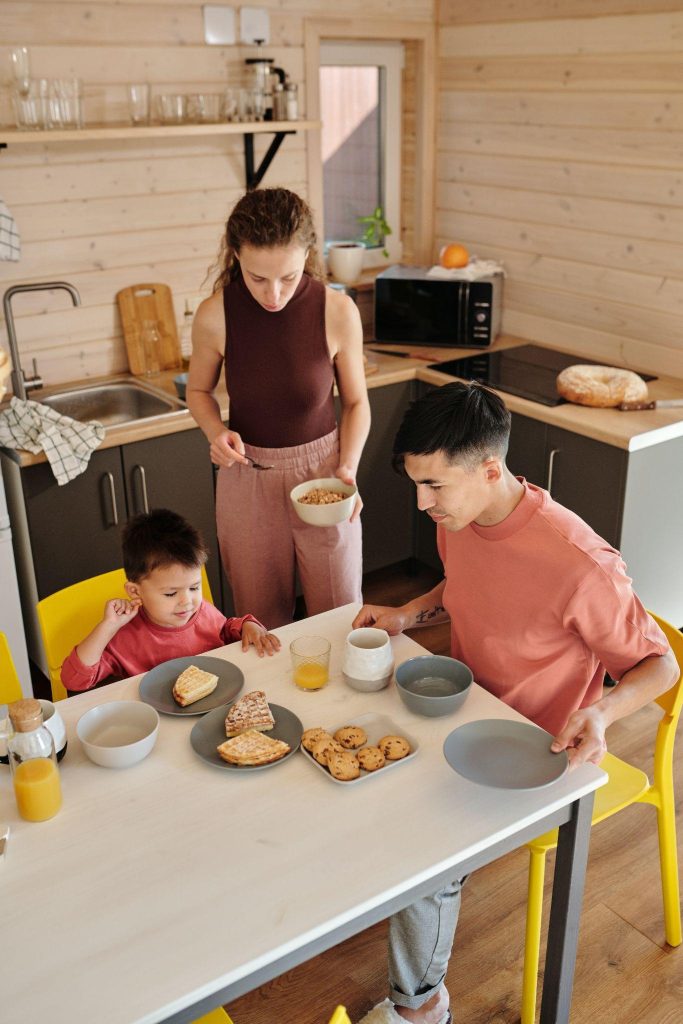 Image of a family of three enjoy a breakfast of cereal, waffles, and muffins.