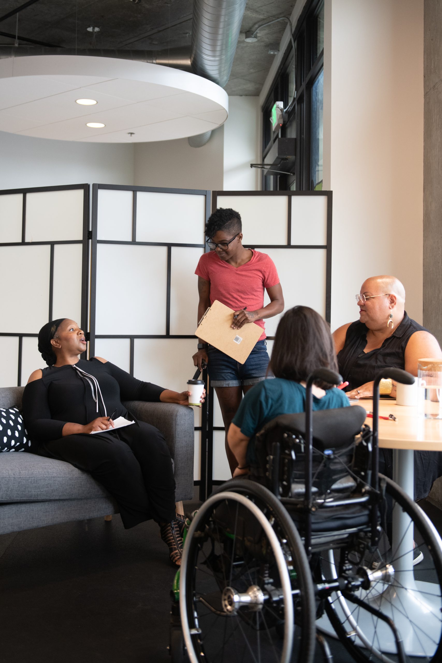 Four disabled people of color gather around a table during a parent-teacher conference. A Black woman sitting on a couch gestures and speaks while the three others (a South Asian person sitting in a wheelchair, a Black non-binary person sitting in a chair, and a Black non-binary person standing with a clipboard and cane) face her and listen.