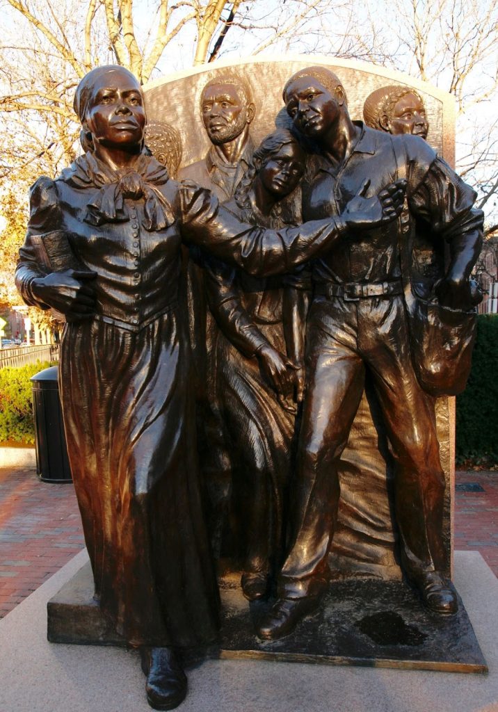 A 10-foot high bronze statue with six Black people; a woman is in front of the others stepping out from the base, with her left arm gesturing to lead them on and her right arm holding a bible.