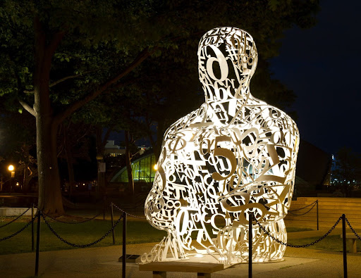 A nighttime photo of a lit white, see-through statue of a human shape made up of different numbers and mathematical symbols