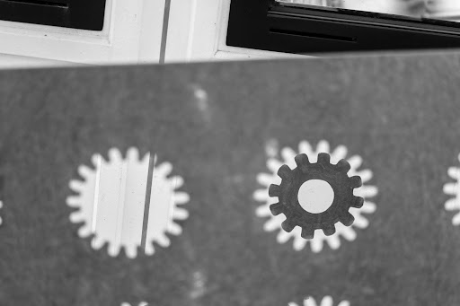 Black and white photo of a drawing of a cog looking through a metal cog-shaped hole