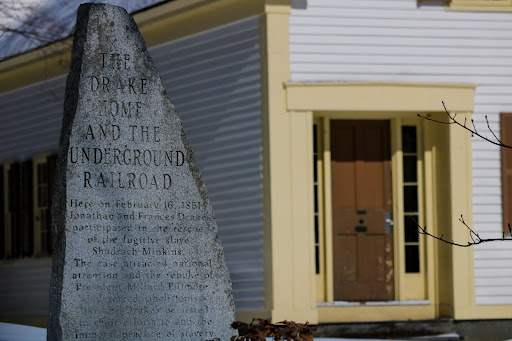 Photo of a white house with yellow trim in the background with a triangular shaped stone slab in front engraved with "The Drake Home and the Underground Railroad"