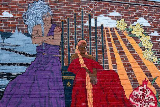 Photo of a mural on a brick wall with a figure on the left with long, white hair and a purple toga and a figure on the right, seated, with dark skin, long red-blonde hair, and a red toga; a large pomegranate on the bottom right