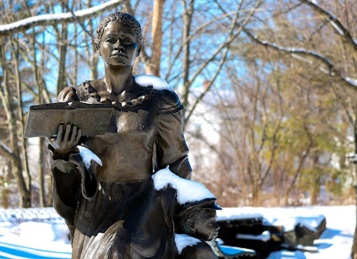 Photo of statue of a female-presenting individual holding a book with arm around a small child, snow on the ground and on the statue