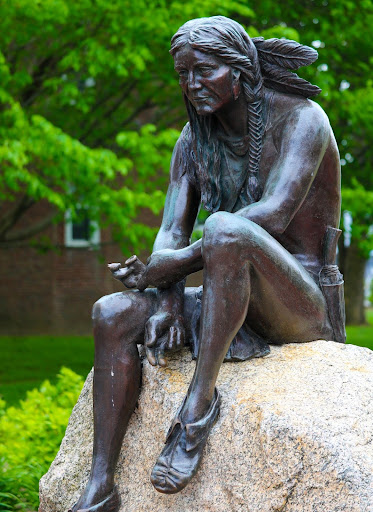 Photo of a statue of a male-presenting Native American sitting on a rock holding a pipe