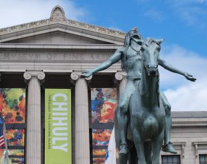 Photo of a statue of a Native American figure on a horse with their head back and arms outstretched in front of a large, columned building