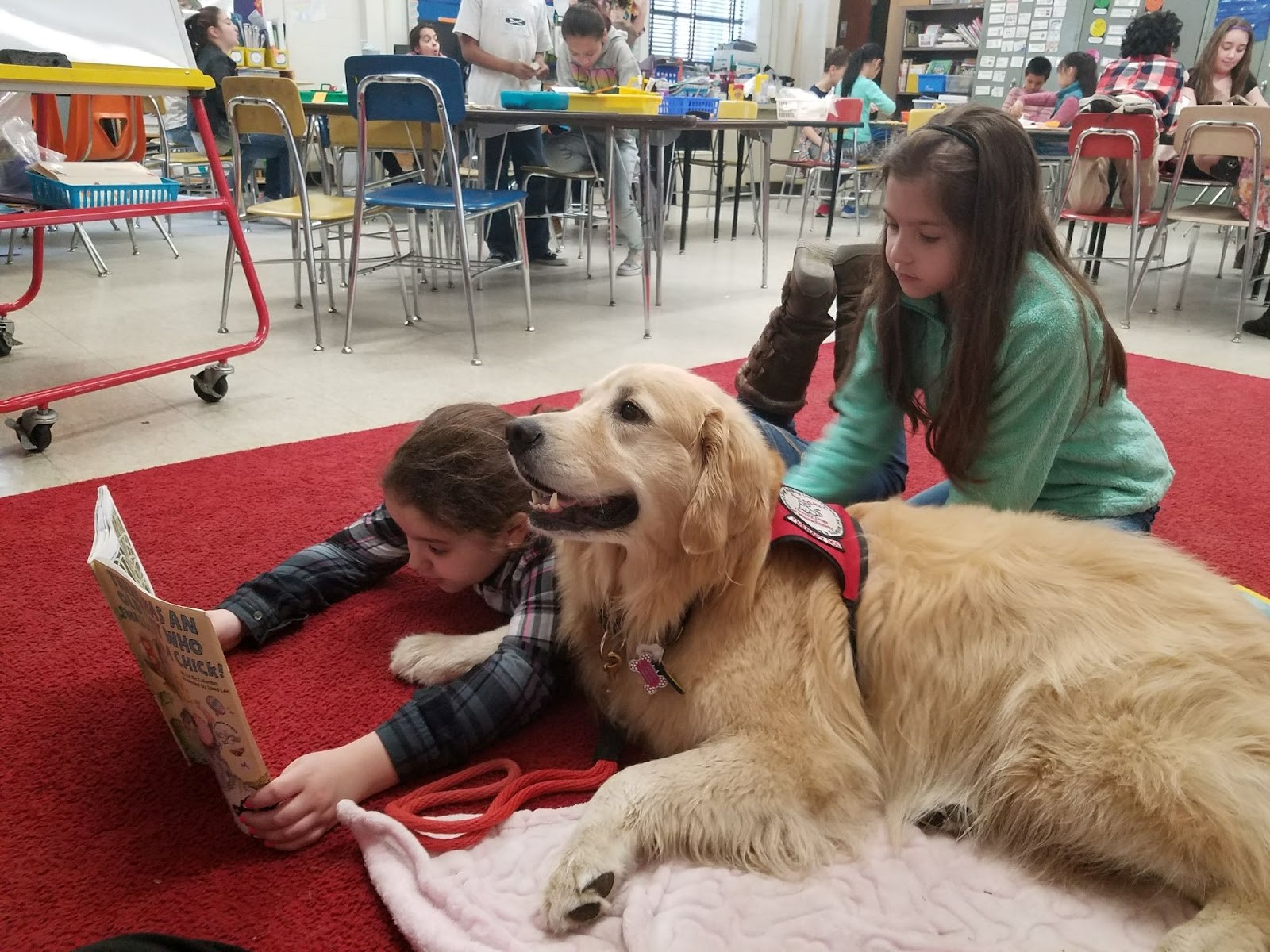 Photo of a dog with a therapy jacket sitting on a red mat with two children, one lying down reading a book